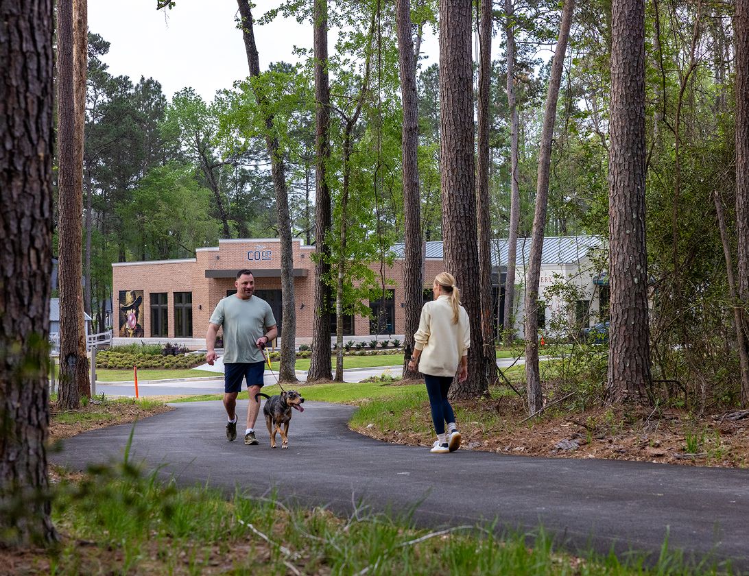 Two people walk on a paved path through a wooded area, one walking a dog, with a modern brick building—typical of new homes in Montgomery County, TX—visible in the background.