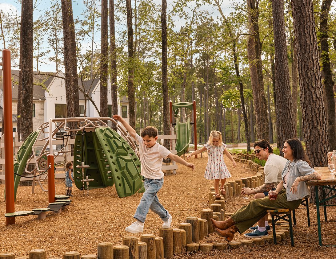 Two children balance on wooden stumps at a playground near The Grazery restaurant at Two Step Farm, while two adults sit at a table. Playground equipment and trees are visible in the background.