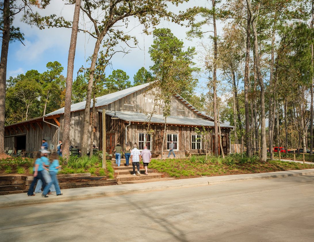 A group of people walk toward and enter The Grazery restaurant at Two Step Farm, a rustic wooden building surrounded by tall trees on a sunny TX day.