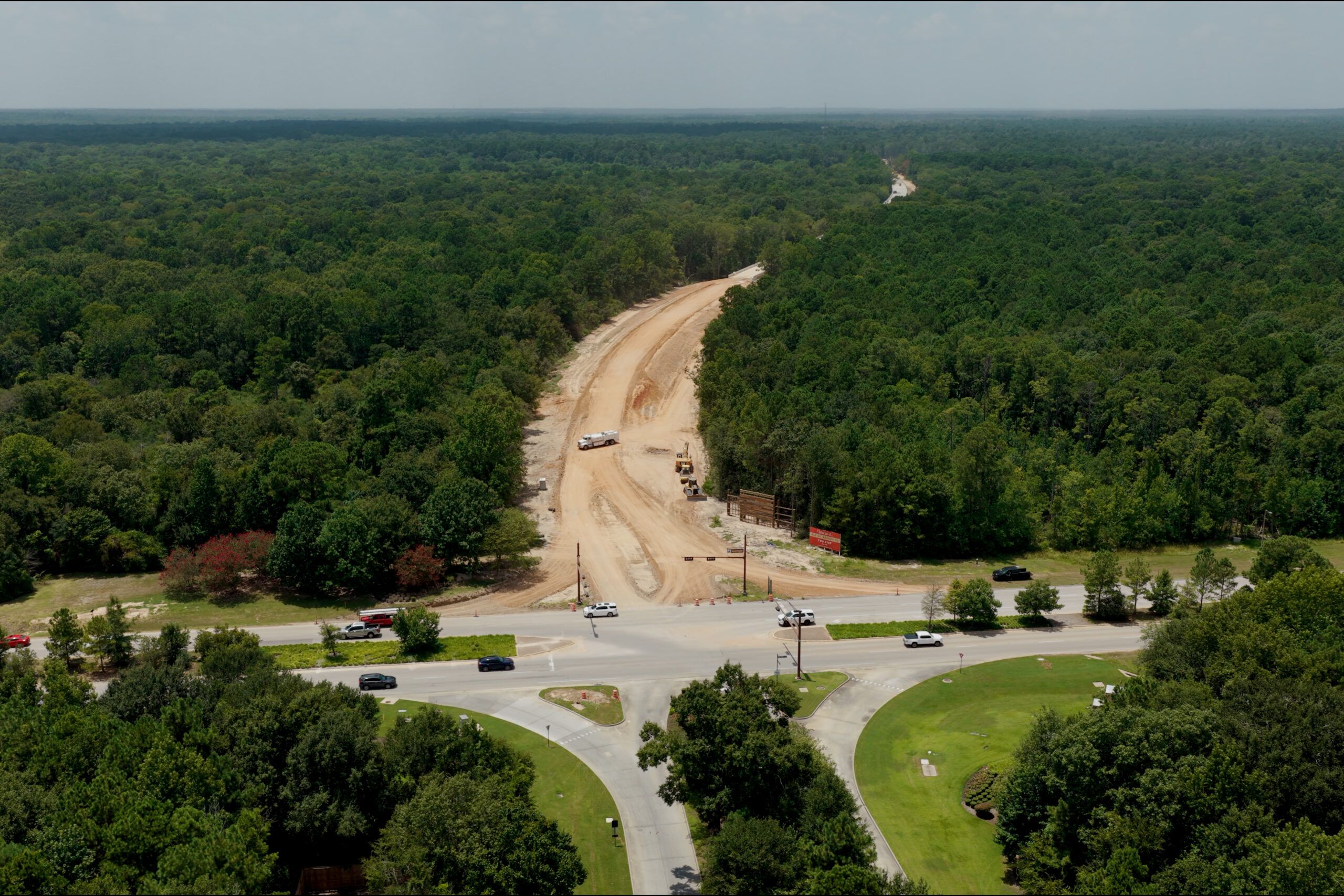 Aerial view of a road construction site cutting through a forest, with vehicles and equipment visible near a major intersection in the foreground.