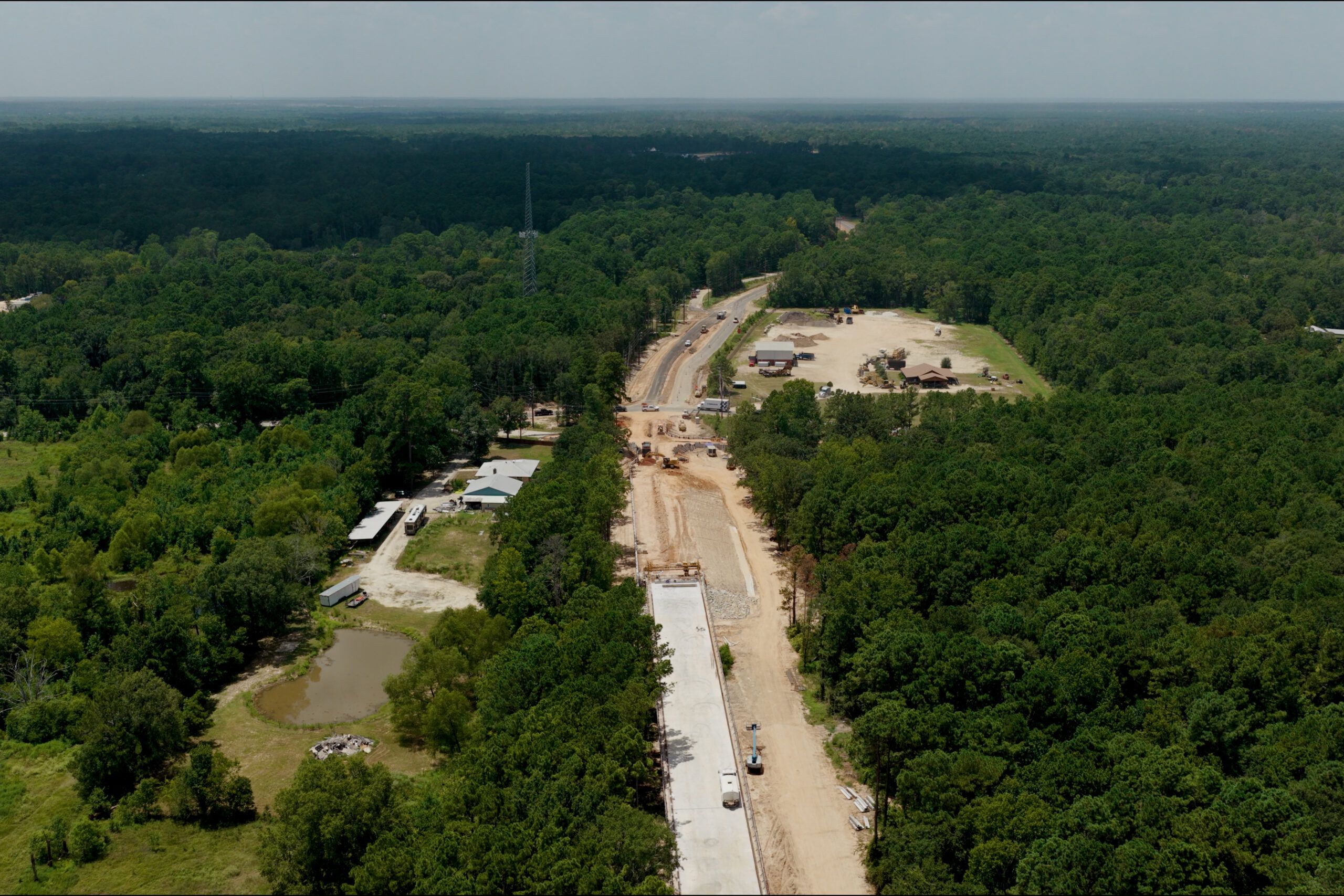Aerial view of a construction site in a forested area, with a partially completed road, vehicles, and several buildings surrounded by dense green trees.