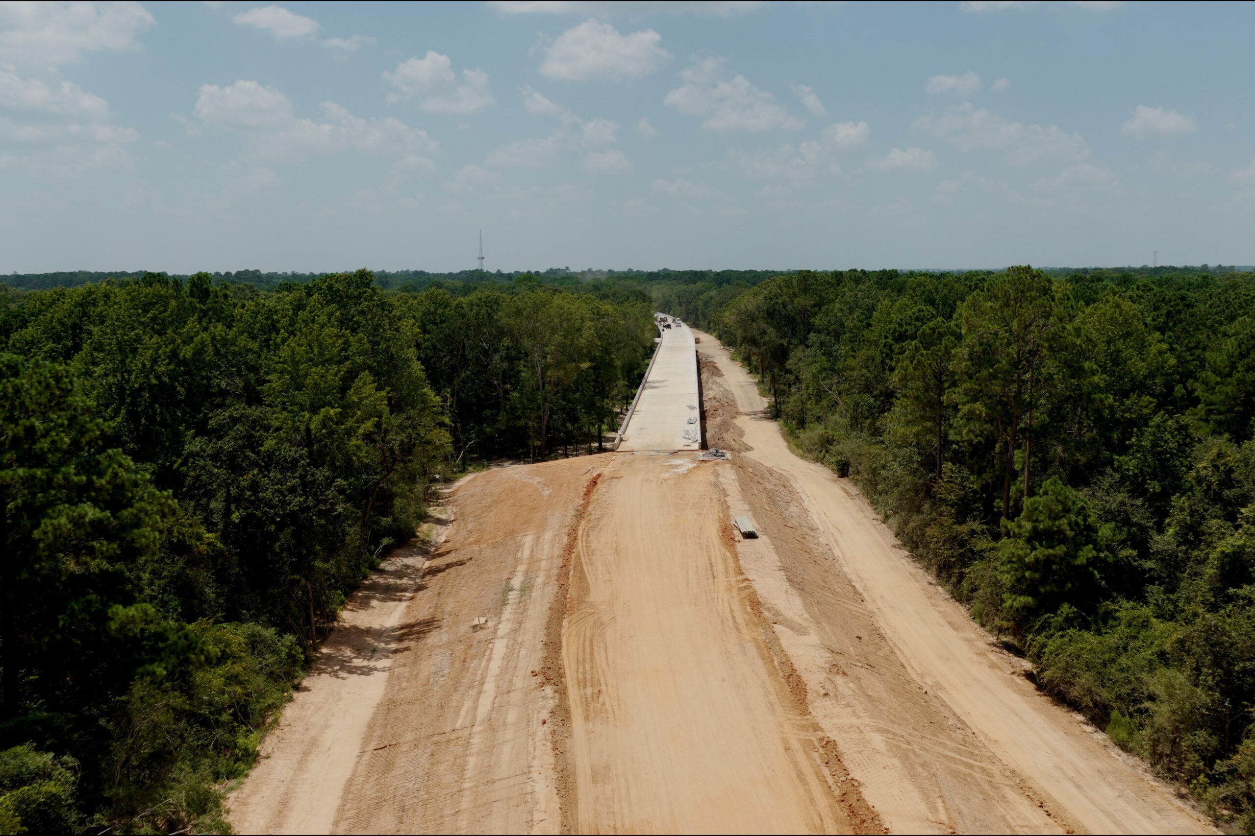Aerial view of a partially constructed road with a paved section in the center, surrounded by cleared dirt and dense forest on both sides under a partly cloudy sky.