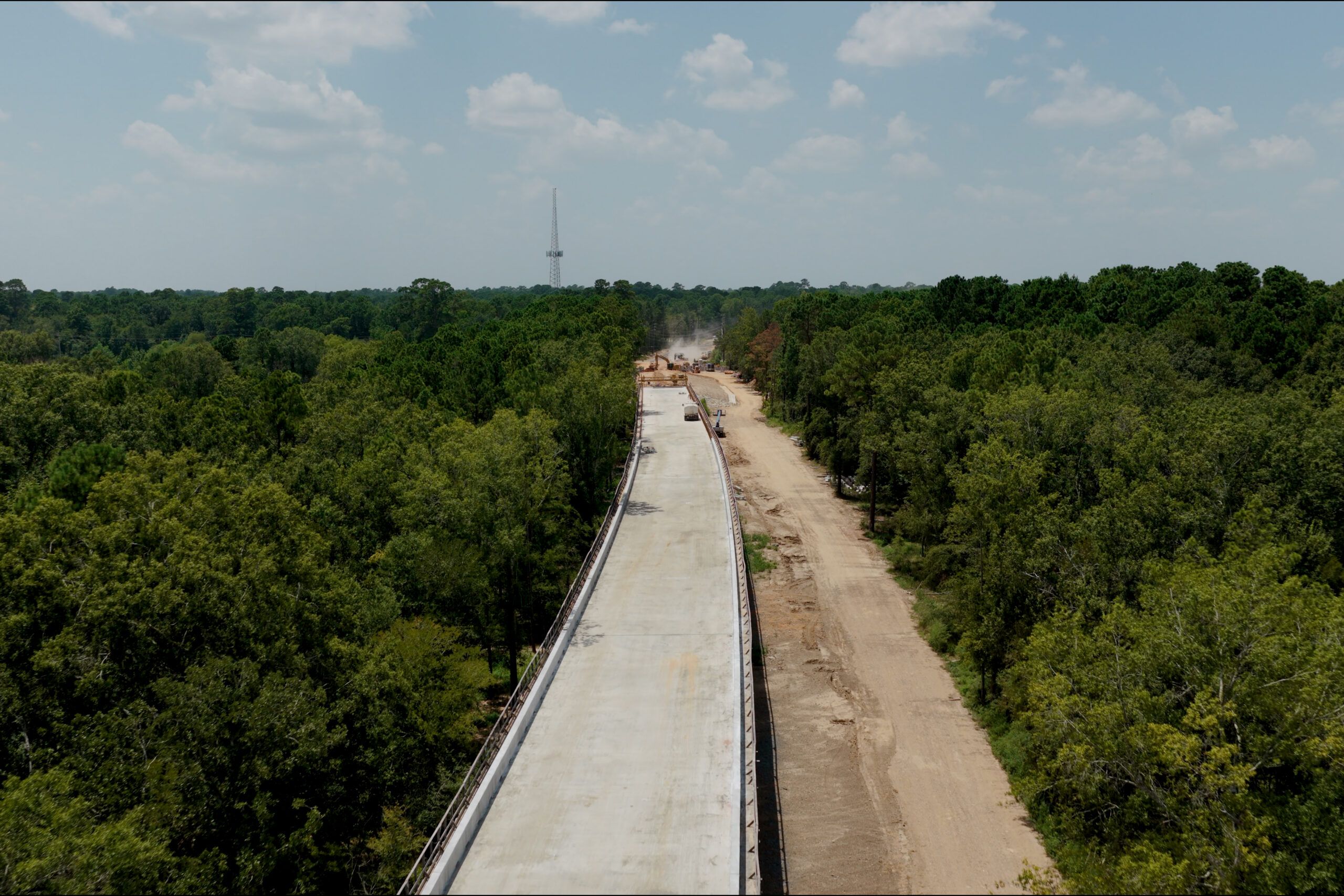 Aerial view of a partially constructed concrete road surrounded by dense green trees, with construction vehicles and equipment visible in the background.