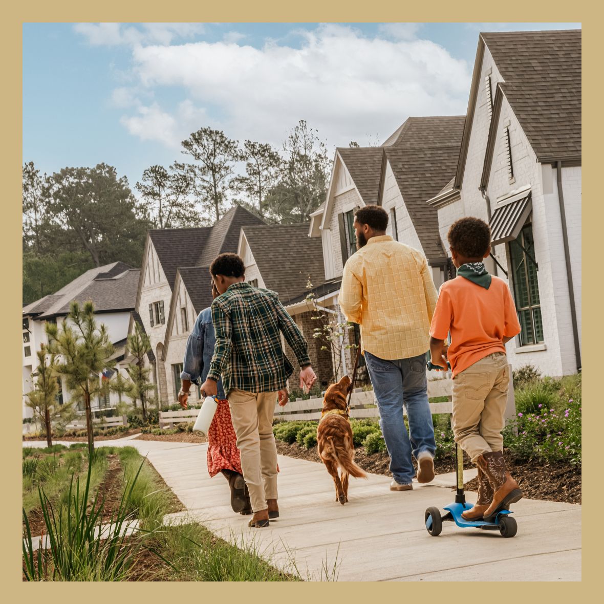 A family of four and a dog walk on a sidewalk in a suburban neighborhood with modern houses and greenery. One child rides a scooter.